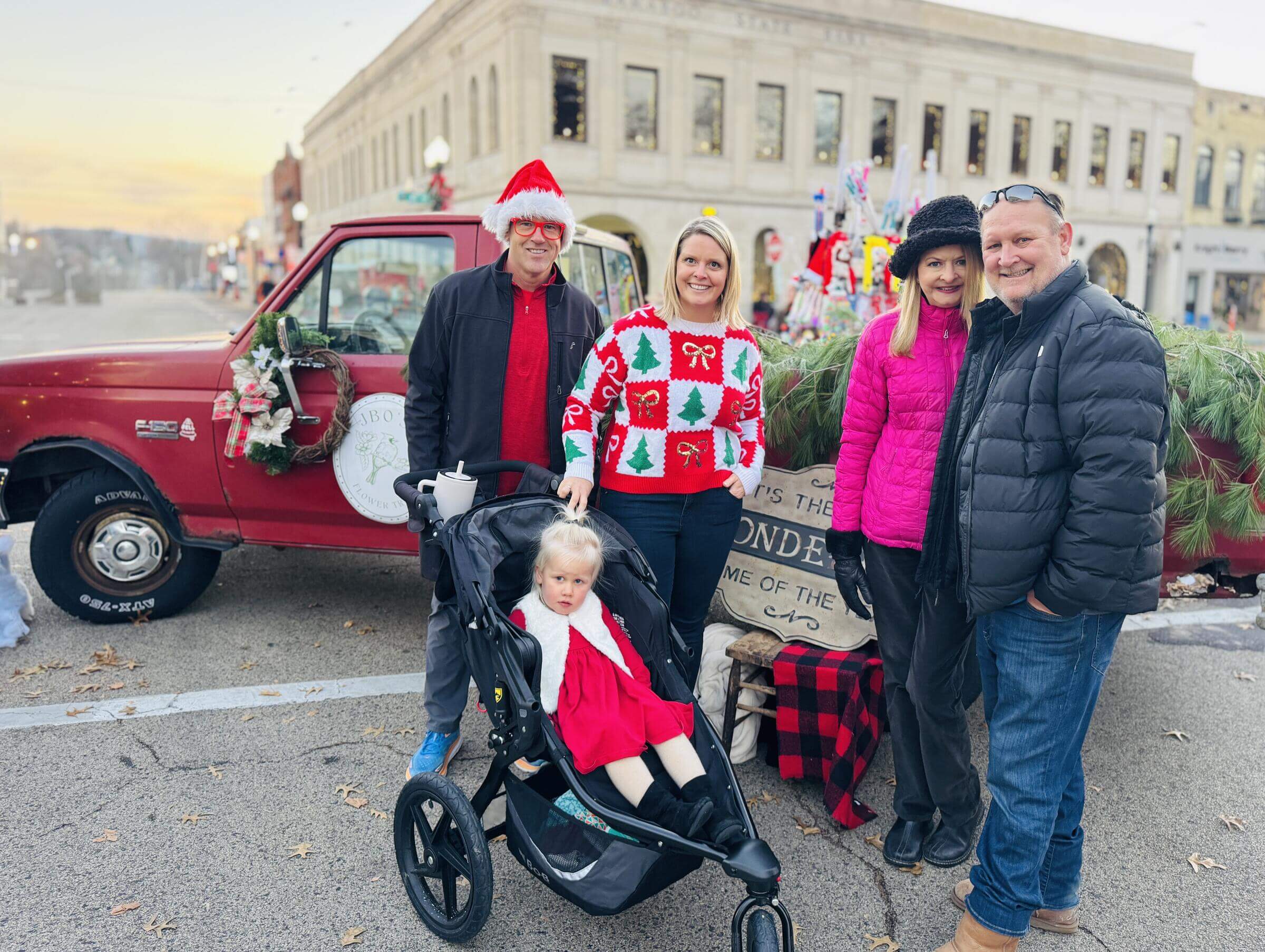 Two adult couples and a child in a stroller. Wearing winter coats by a red truck and Christmas decorations
