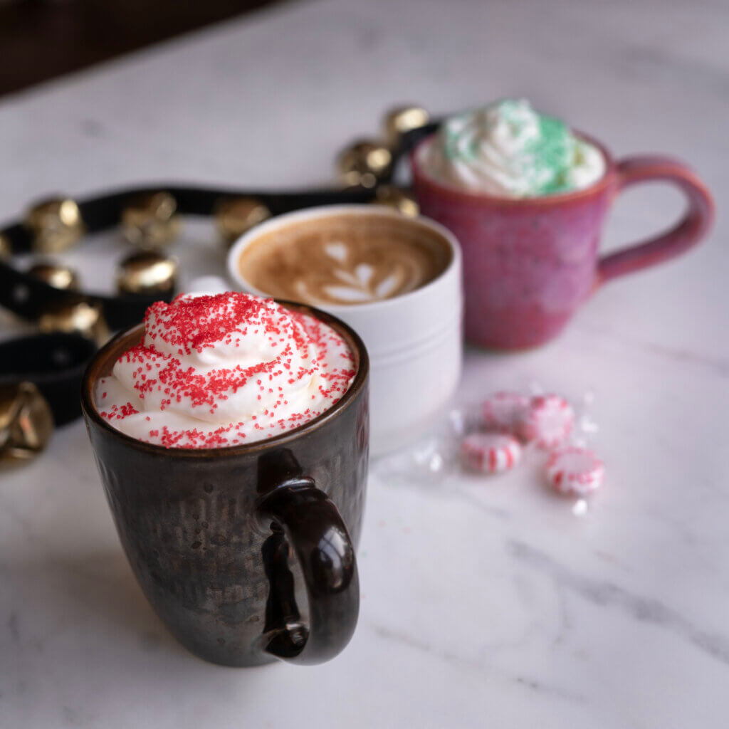Three holiday drinks lined up on a counter. Hot chocolate and coffee drinks with whipped cream and sprinkles